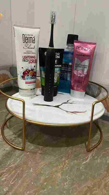 Cosmetic products including a bottle, scrub, and toothbrush on a marble tray with gold frame.